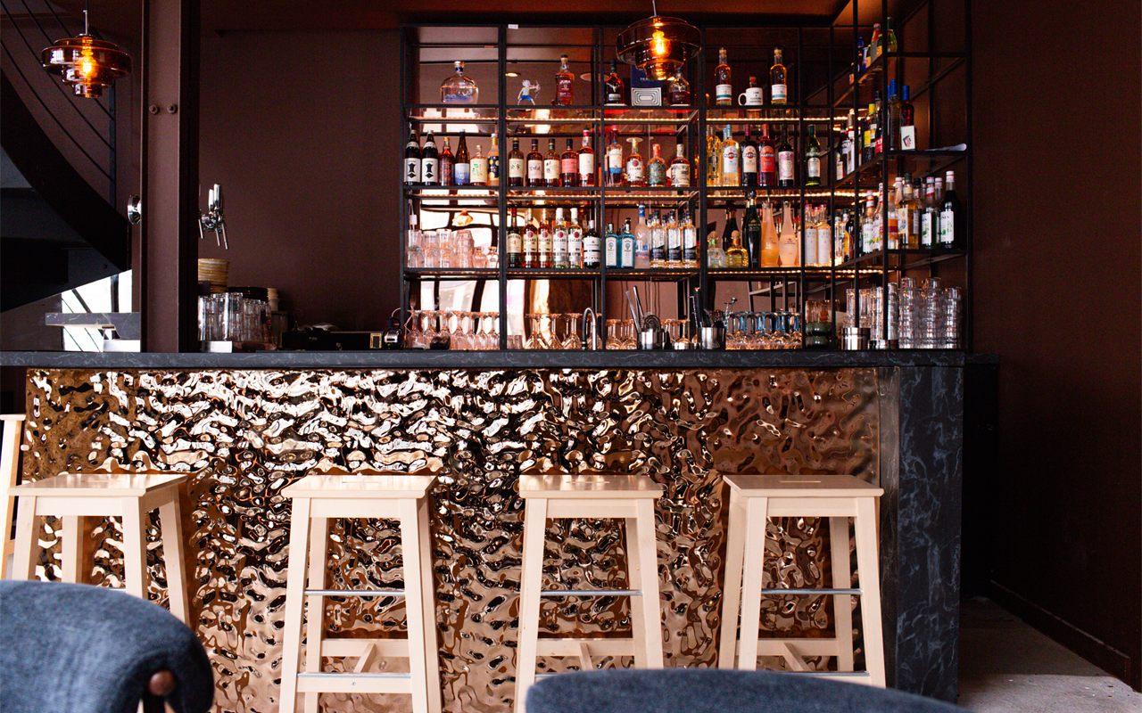 Le bar de Suki Paris, façade en cuivre martelé, tabourets en bois et étagères de bouteilles