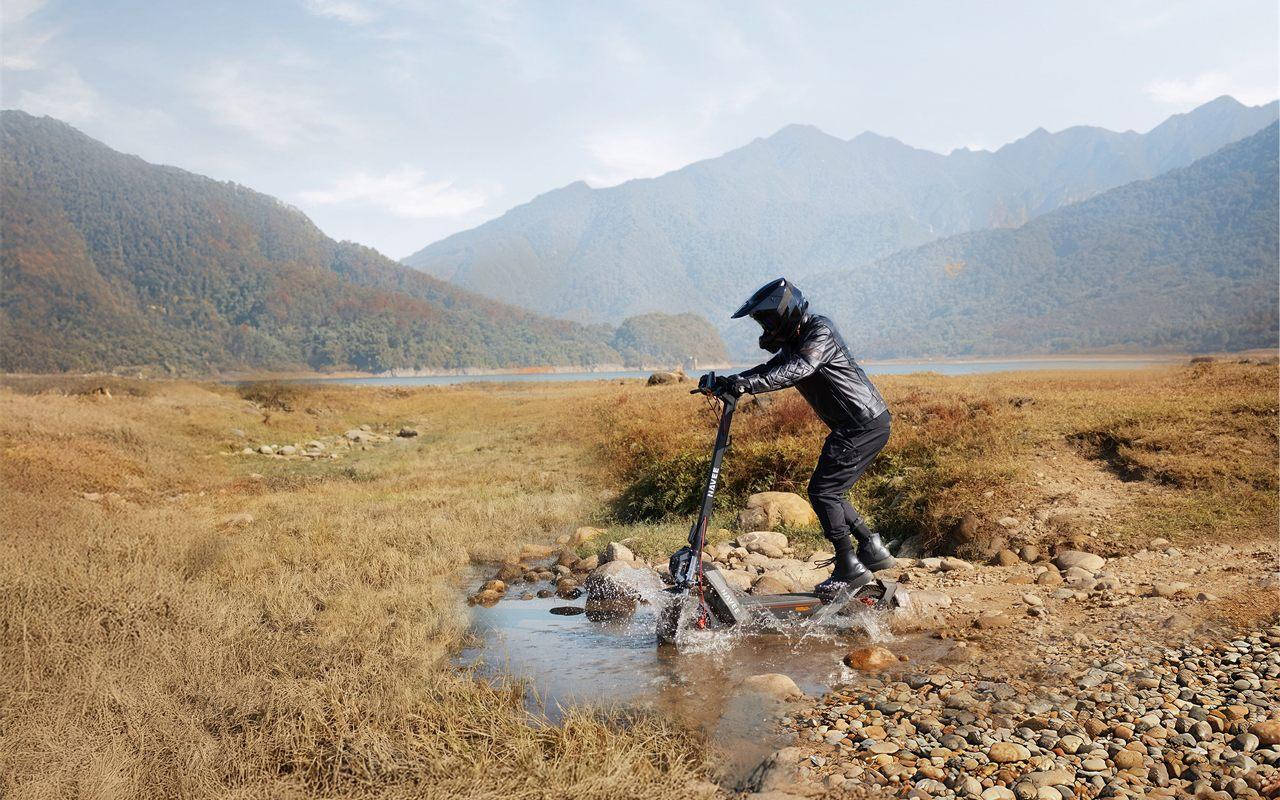 La NAVEE NT5 Max traversant un cours d'eau peu profond, gerbes d'eau autour des roues