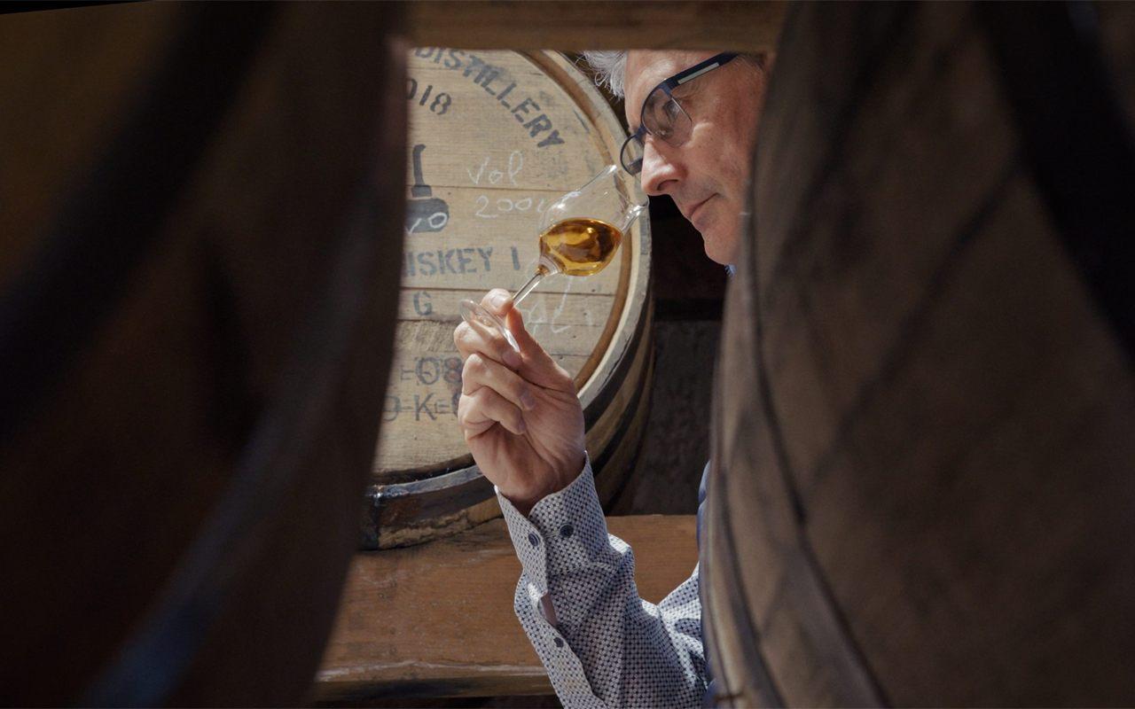 Un verre de single malt ABK6 levé à la lumière, entre deux fûts de chêne