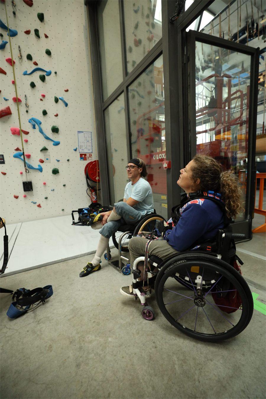 Deux grimpeurs en fauteuil roulant, baudrier enfilé, au pied d'un mur d'escalade