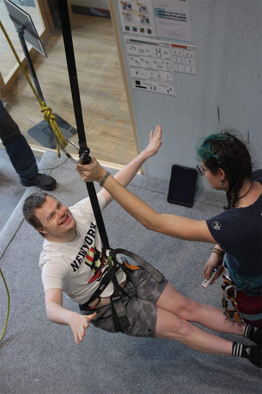 Un grimpeur assis dans son baudrier, soulevé par le Footing Vertical, sourit à la monitrice qui l'accompagne