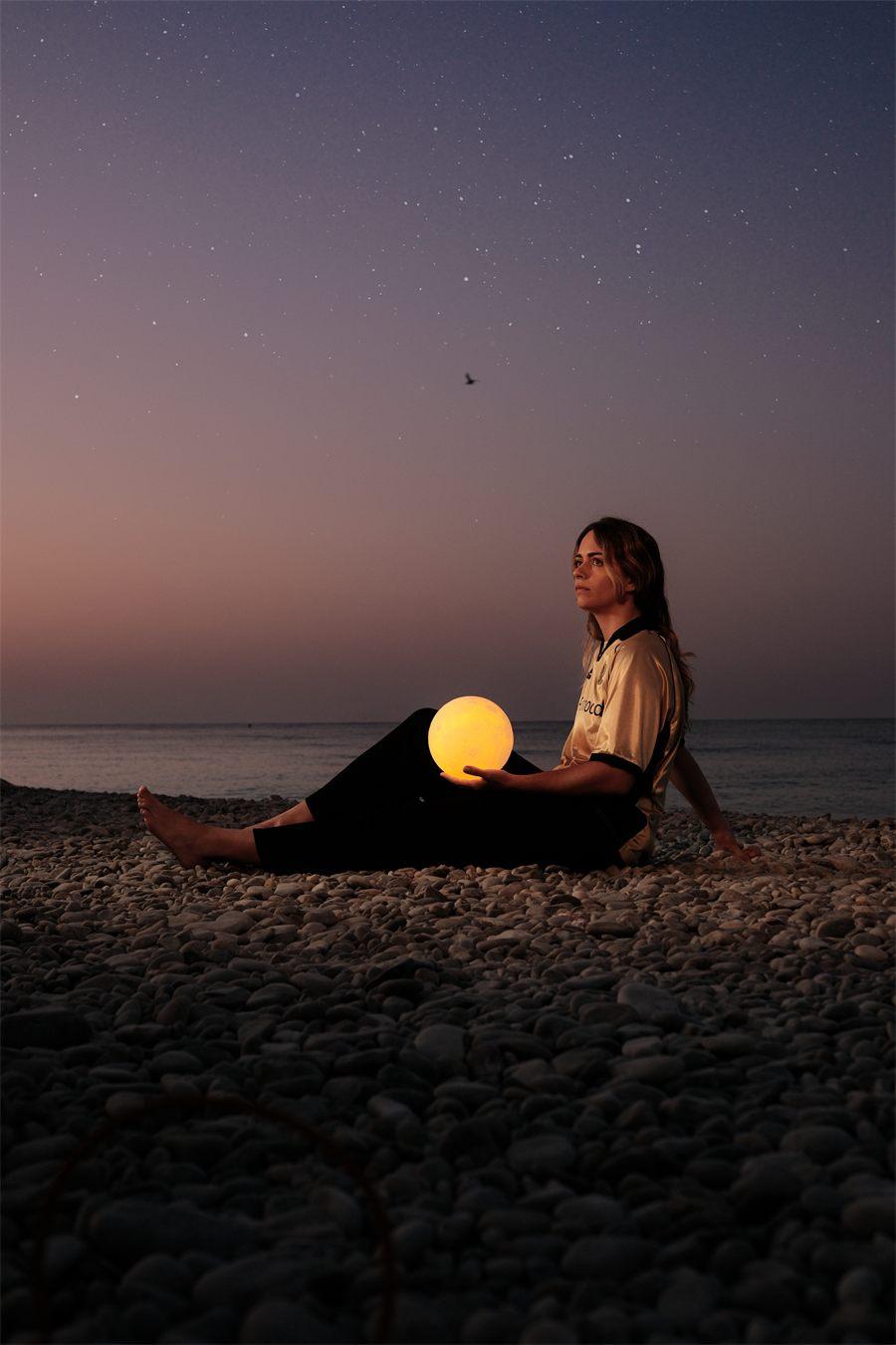 Assise sur les galets face à la mer, une supportrice tient une boule lumineuse à l'aube