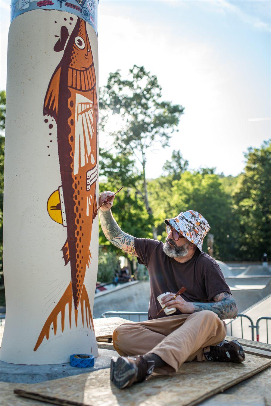 Jeremy Fish assis sur la plateforme, en train de peindre le poisson sur la colonne du Volcano Opinel