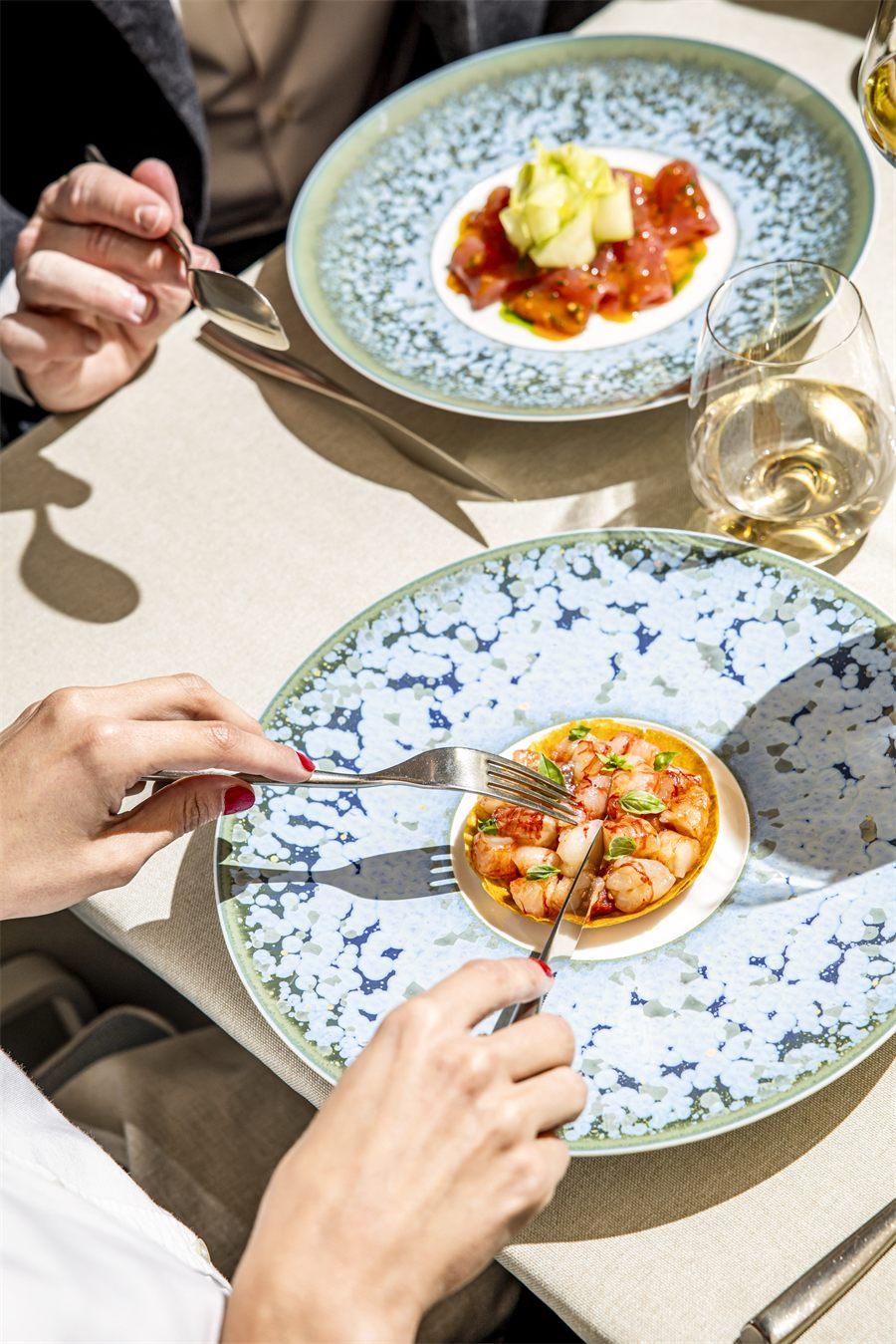 Deux convives déjeunent en terrasse au Pavyllon Paris, couverts en main devant des assiettes en céramique bleue et un verre de vin blanc