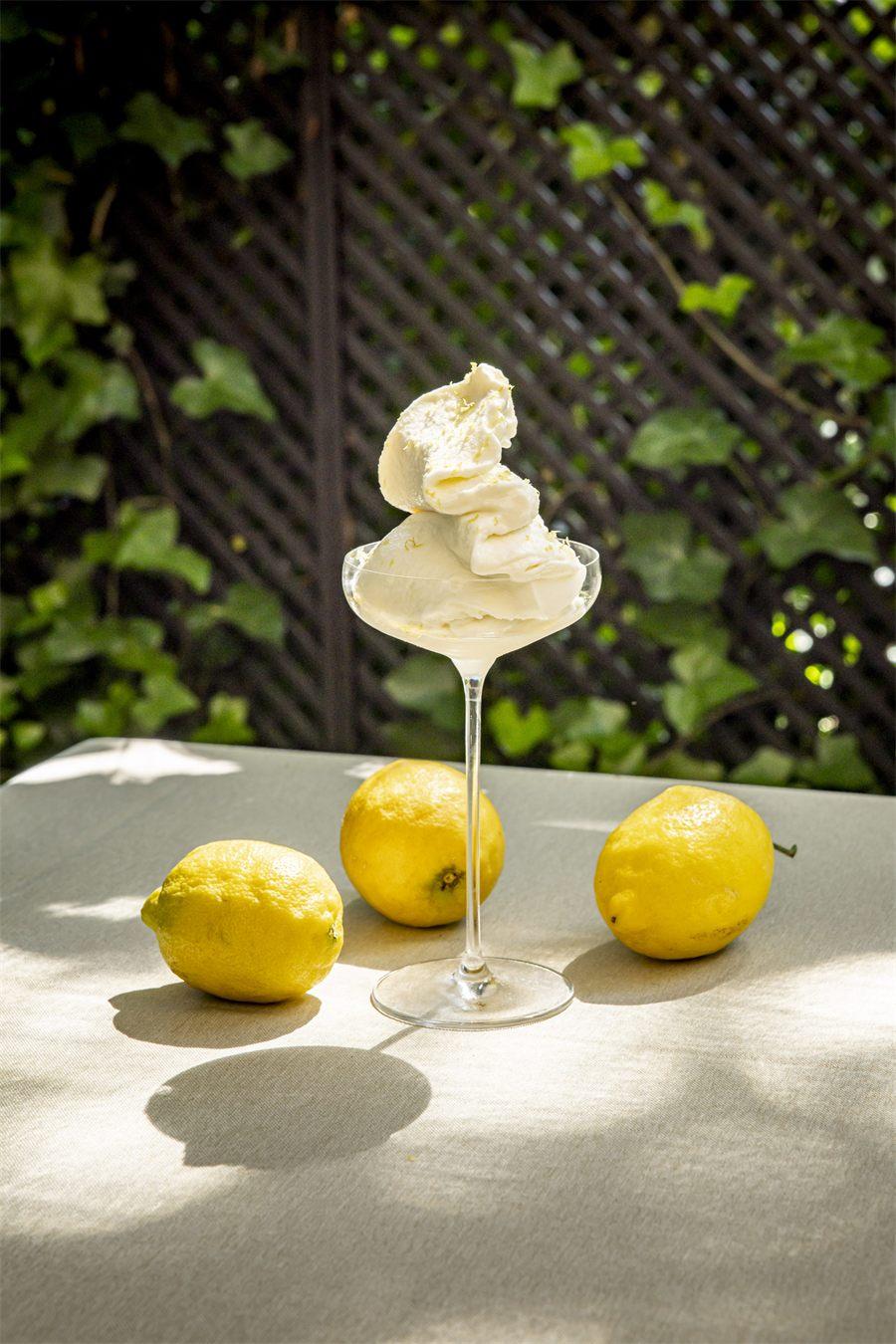 Coupe de sorbet au citron parsemée de zestes, trois citrons entiers posés sur la nappe, treillage couvert de lierre en arrière-plan