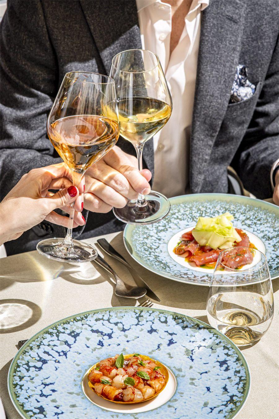 Deux convives trinquent avec un verre de vin blanc et un verre de vin orange au-dessus de deux assiettes en céramique bleue, en terrasse au soleil