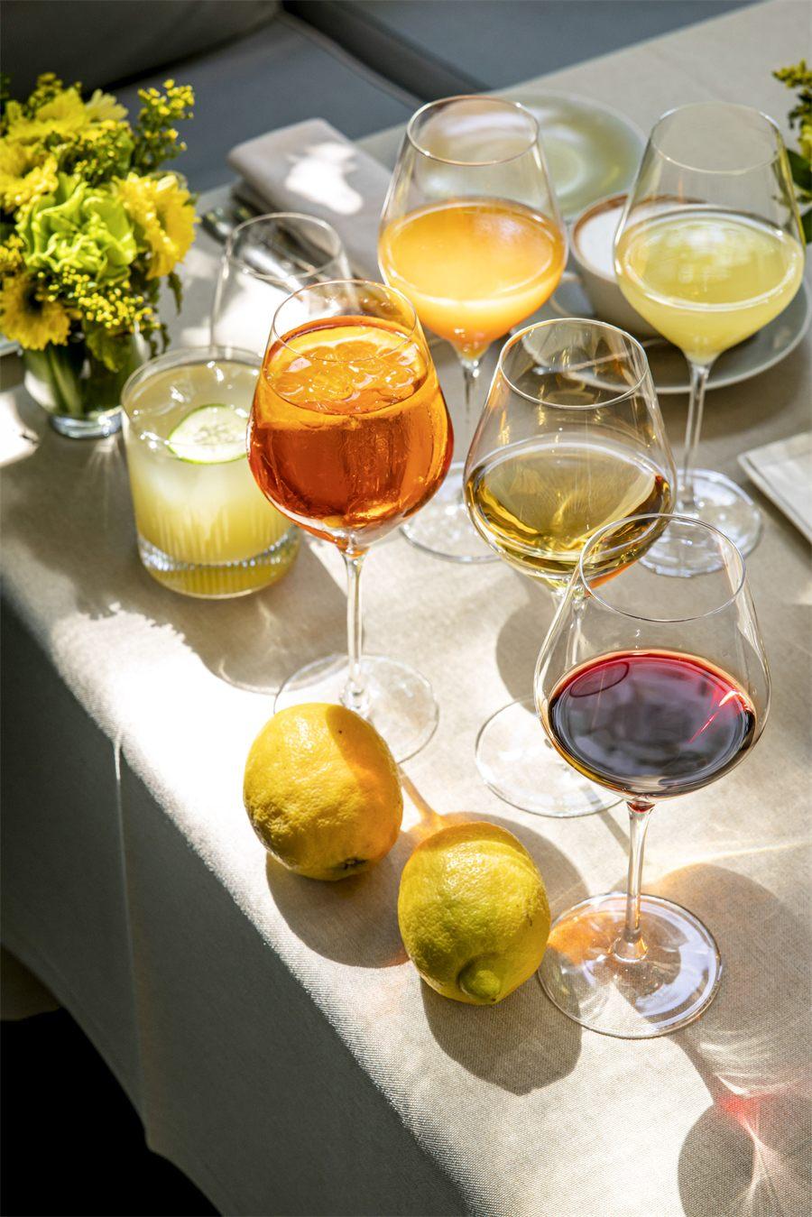 Verres de vin blanc et orange, spritz et cocktail au concombre posés sur une nappe claire, entre citrons et bouquet de fleurs jaunes