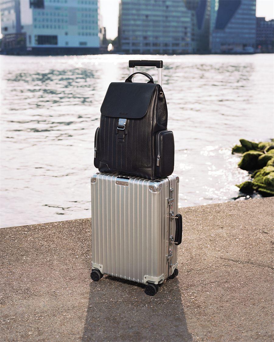 Sac a dos Never Still en cuir noir accroche par son passant arriere a la poignee telescopique d une valise RIMOWA