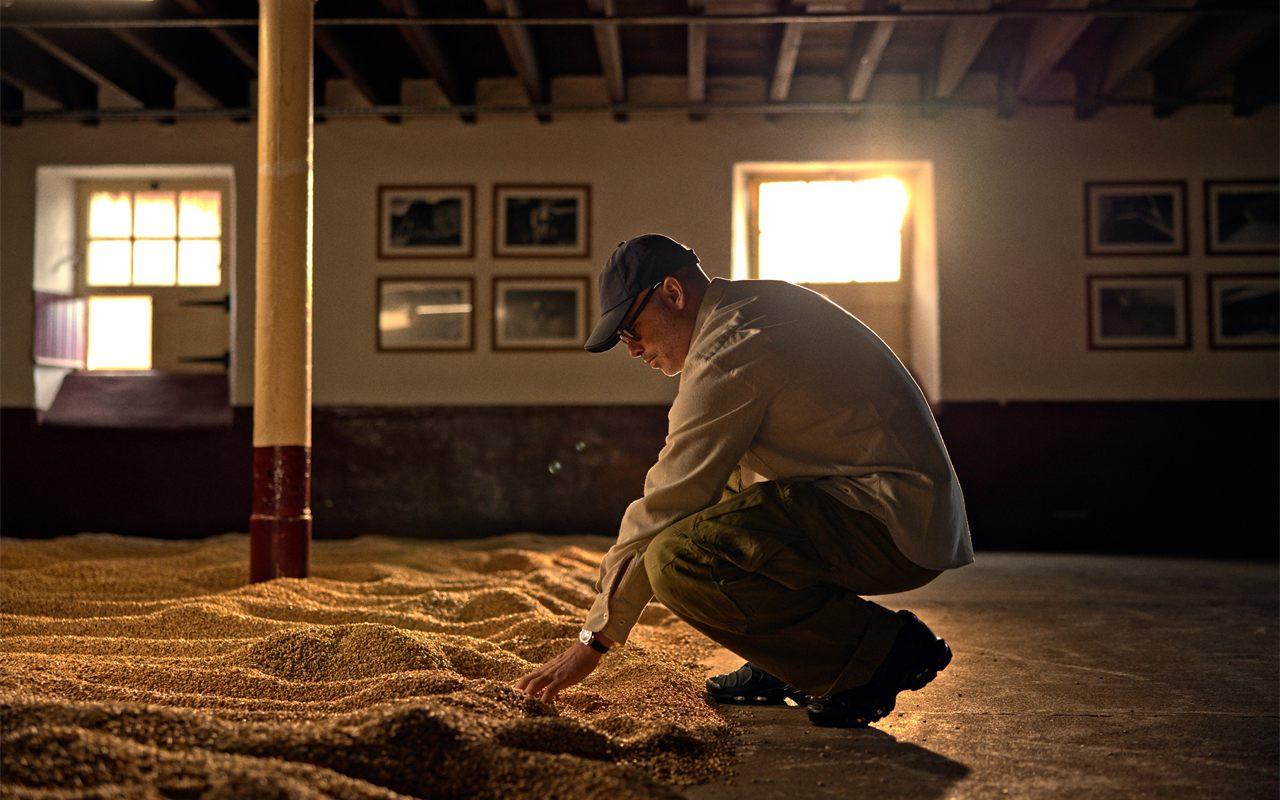 Daniel Arsham accroupi sur l'aire de maltage de The Balvenie, la main dans l'orge