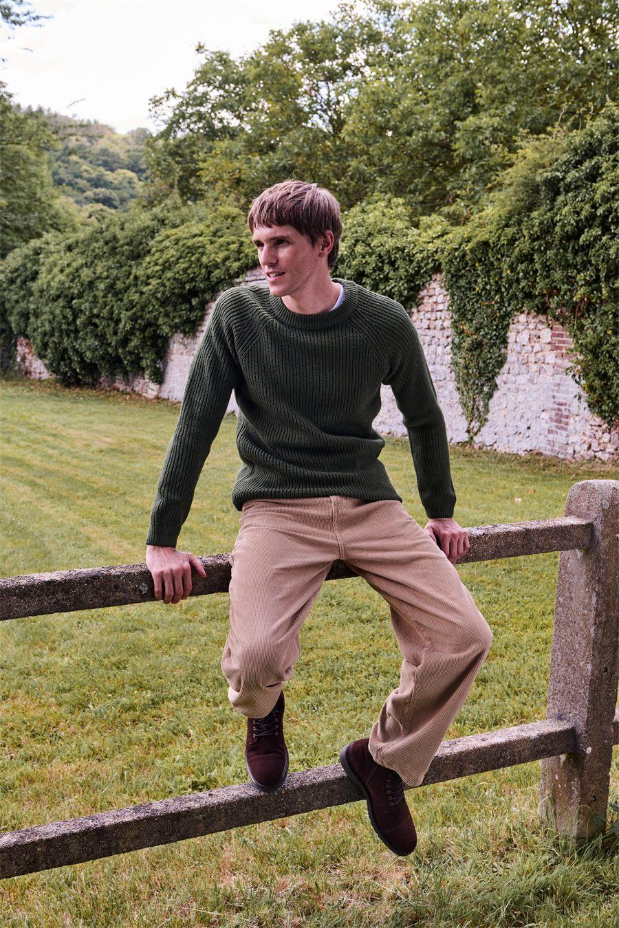 Homme assis sur une clôture en bois, pull en grosse laine et boots Samuel de Bocage