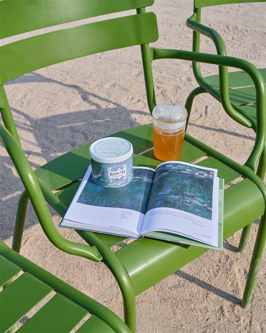 Boîte Kusmi Tea Les Nymphéas posée sur un livre sur les Nymphéas, sur une chaise verte, à côté d'un verre de thé