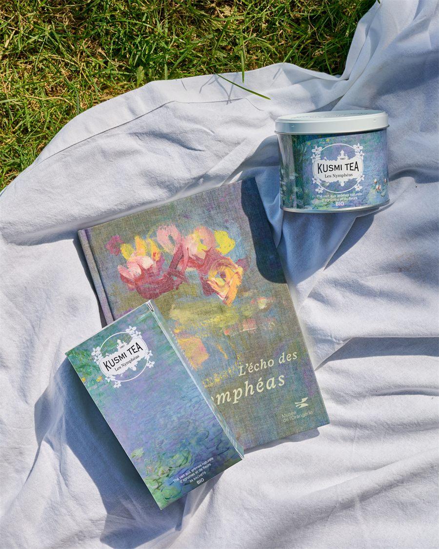Boîte de 100 g et boîte de 24 sachets du thé Kusmi Tea Les Nymphéas posées sur un livre, sur une nappe dans l'herbe