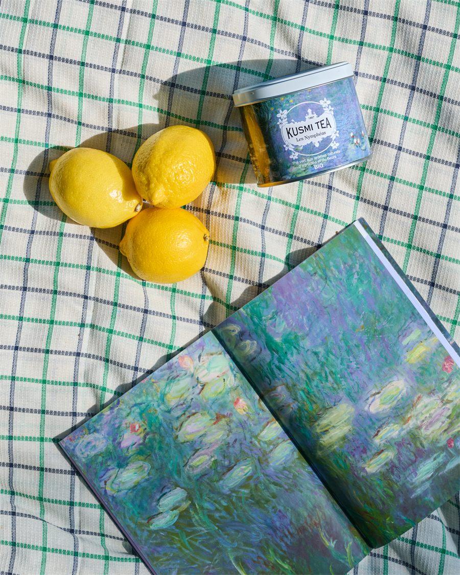 Boîte Kusmi Tea Les Nymphéas, trois citrons et un livre ouvert sur les Nymphéas posés sur une nappe à carreaux