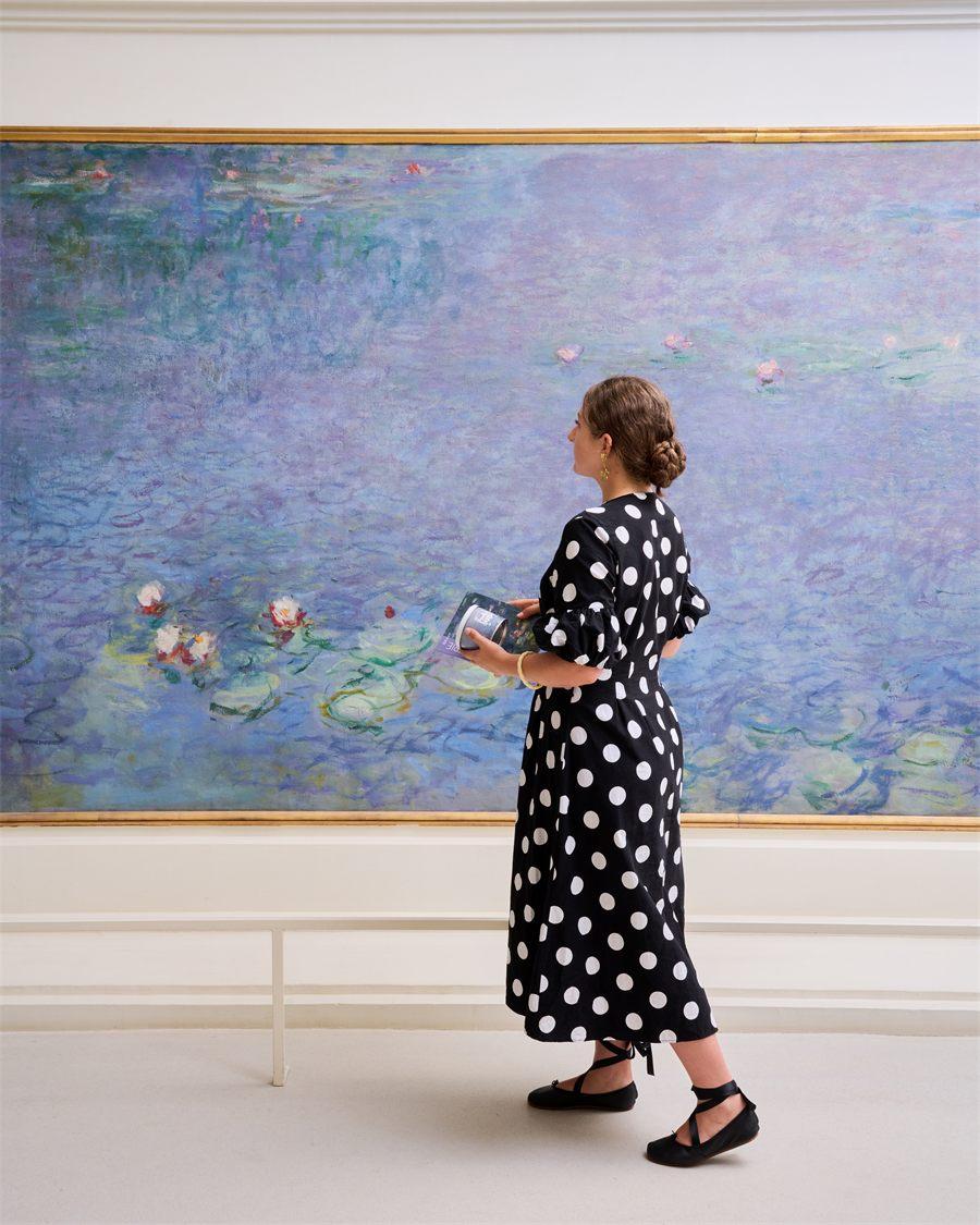 Visiteuse en robe à pois face à un panneau des Nymphéas de Monet, la boîte Kusmi Tea à la main