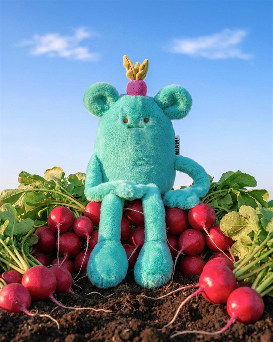 La peluche MIAM Billie Radish assise sur un tas de radis sous un ciel bleu